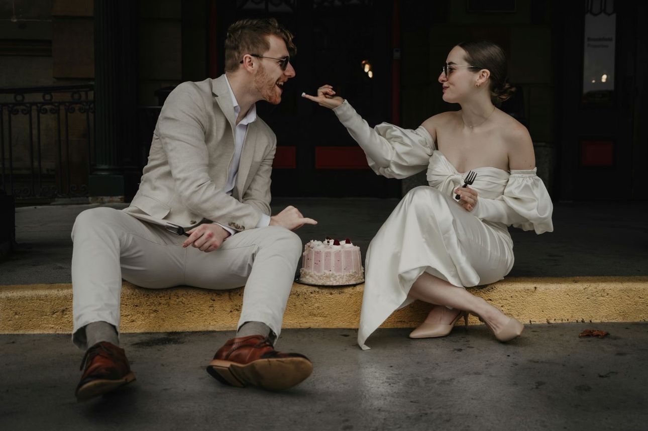 couple in wedding attire sat in road eating wedding cake